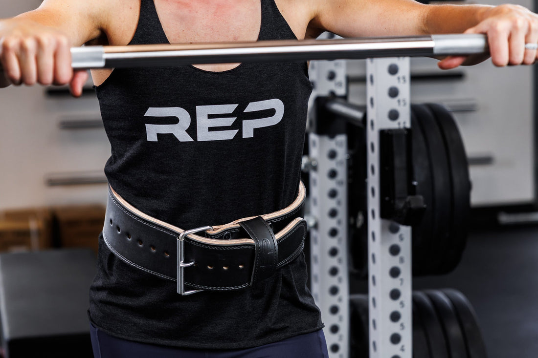 An athlete in a REP® tank top steps up to a barbell with their lifting belt strapped on.